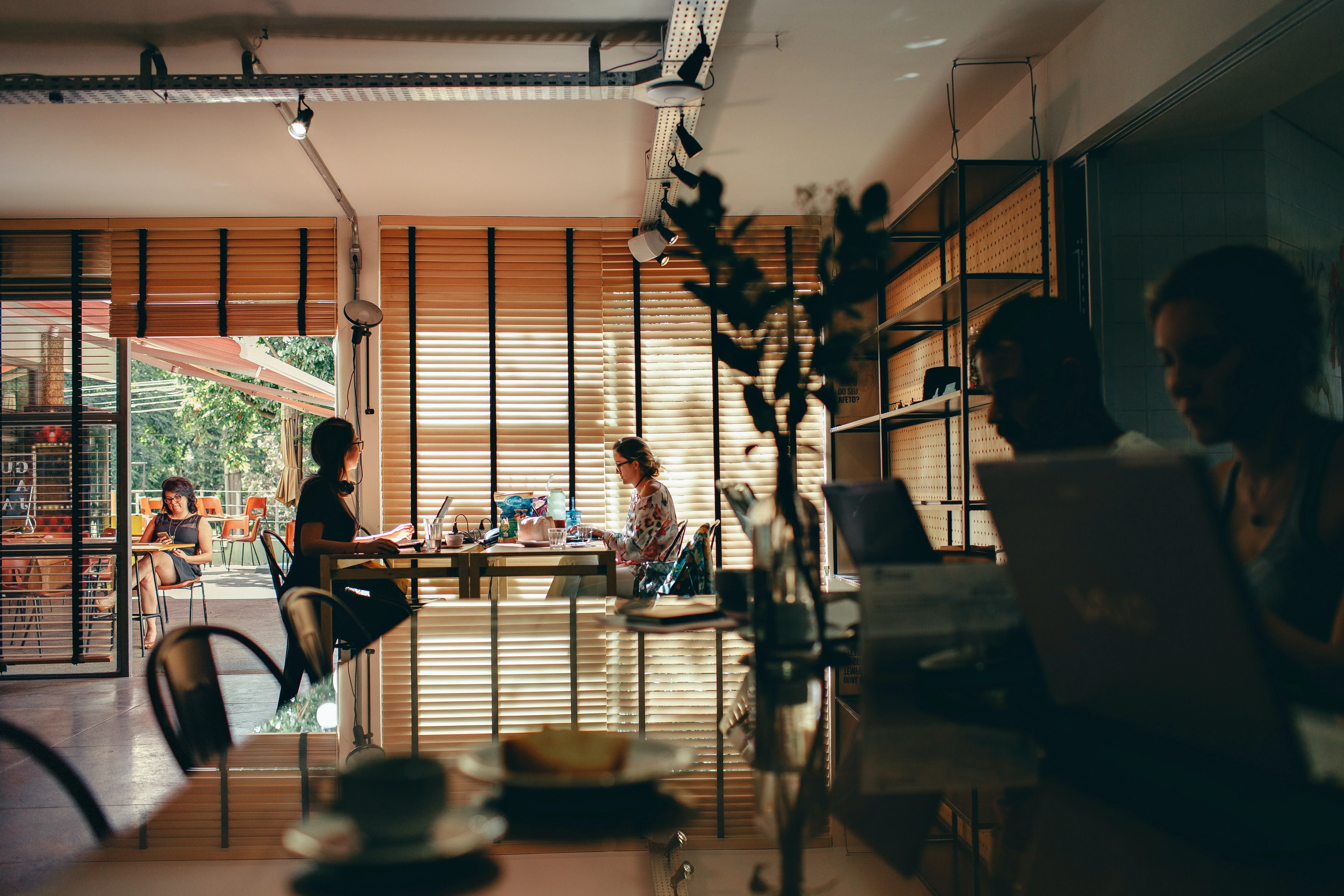 A cozy cafe scene with several people working on laptops, sunlight filtering through wooden blinds, and a modern interior design featuring tables and chairs.