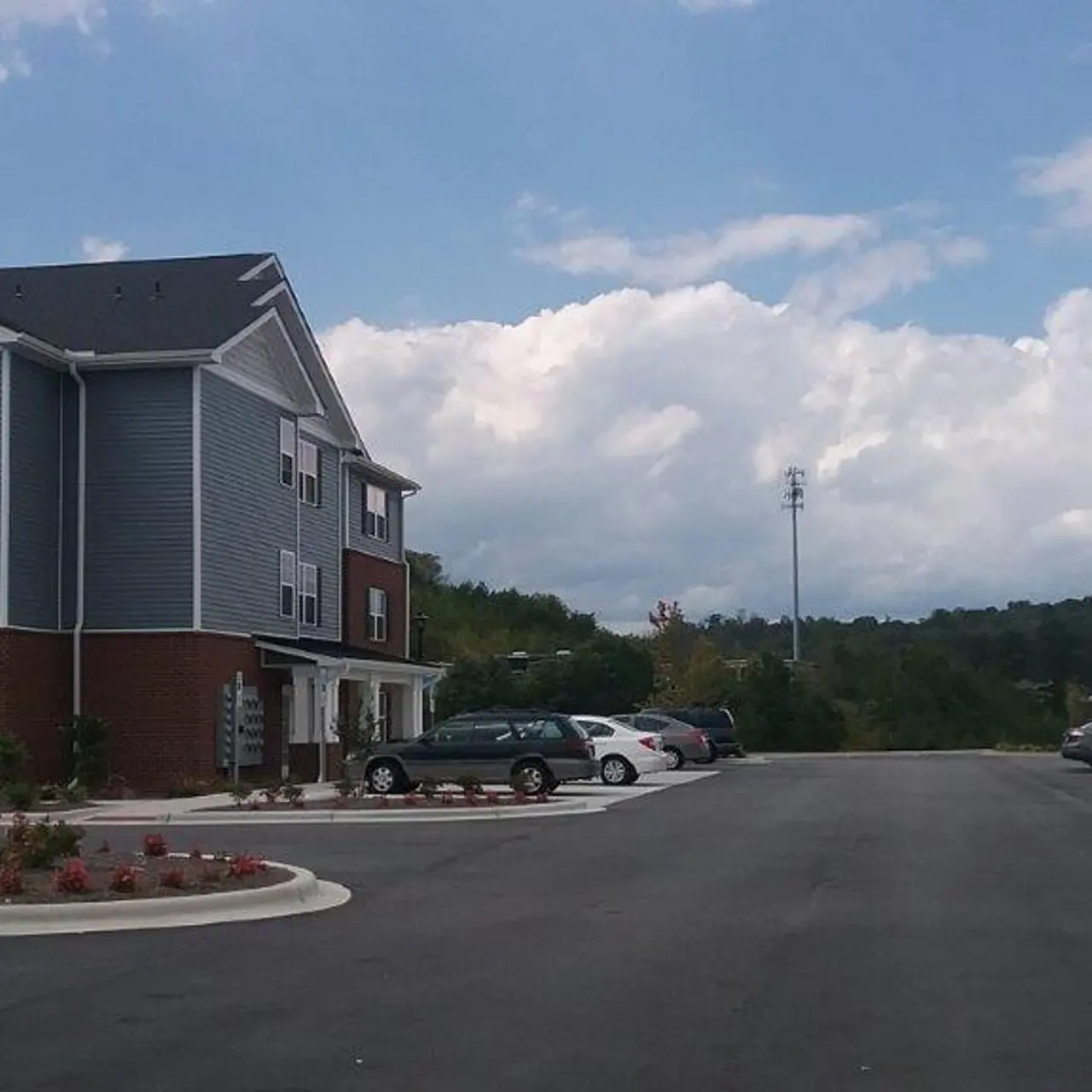 An image showing an apartment complex with a parking lot and several cars parked. The building features a mix of brick and siding, with multiple windows, under a partly cloudy sky.