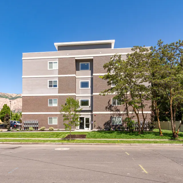 Modern Apartment Building A modern four-story apartment building with a mix of gray and beige exterior, surrounded by trees and greenery, under a clear blue sky.