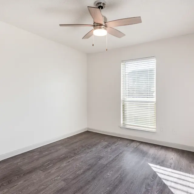 A spacious empty room with a ceiling fan, featuring laminate flooring and a window with blinds.