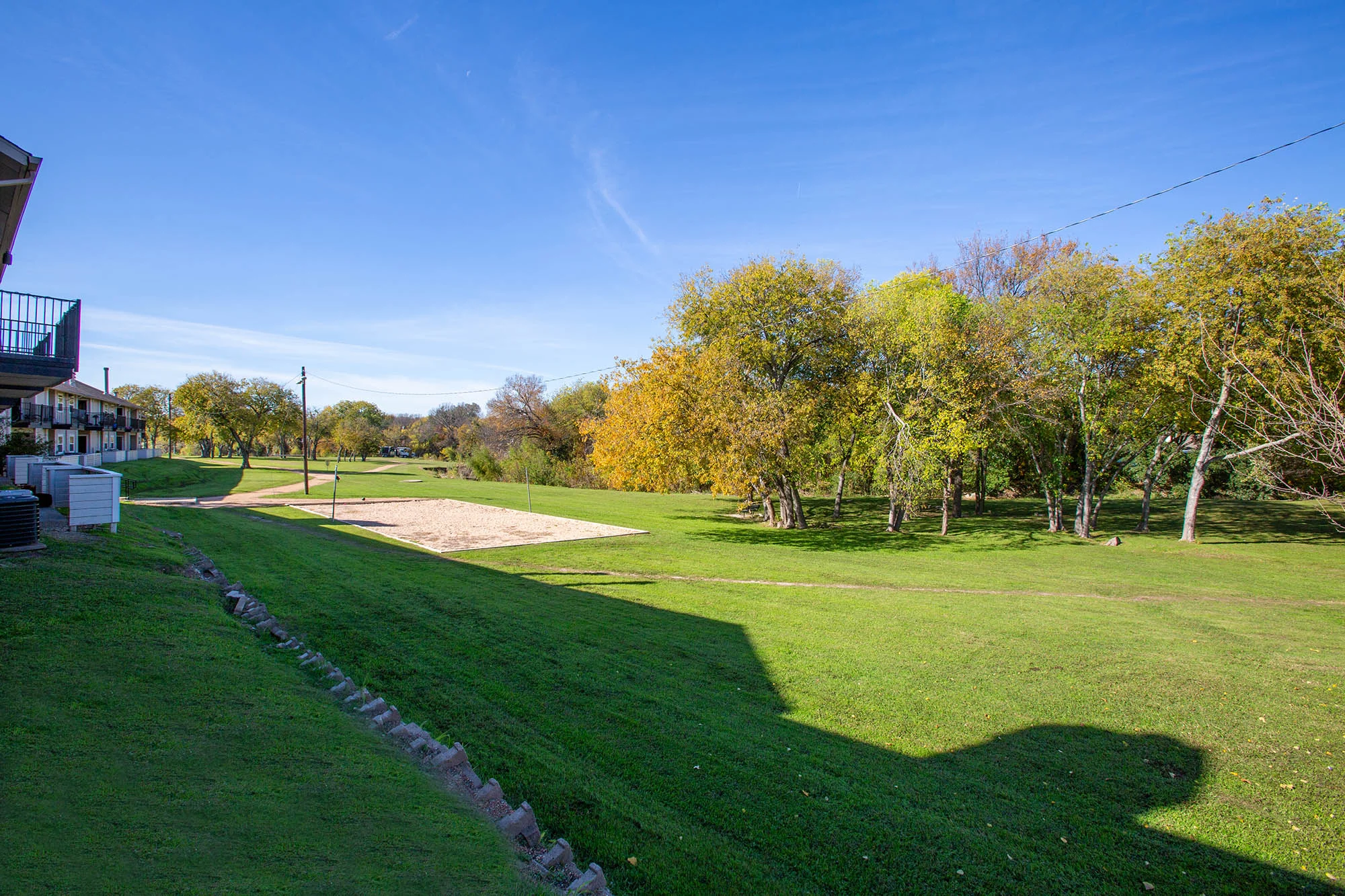 An open green space with a volleyball court, surrounded by trees showing autumn colors and a blue sky.