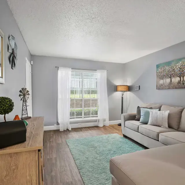 A modern living room featuring a light gray wall, a beige sectional sofa, a wooden side table with a decorative item, and large windows with white curtains. A mint green area rug is on the wooden floor, and a wall art piece depicting trees is hanging above the sofa.