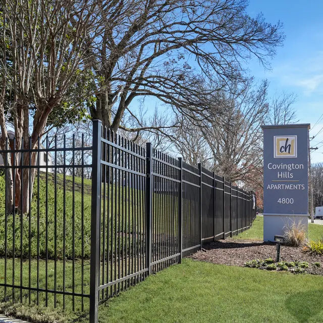 Exterior view of Covington Hills Apartments, featuring a black wrought-iron fence, a sign for the apartments, and well-kept landscaping with a small garden and trees.