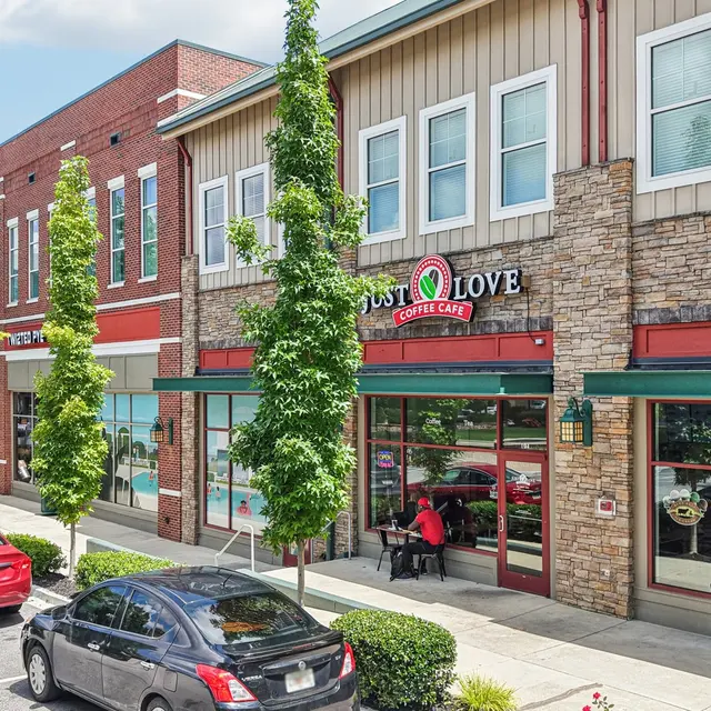A street view of a quaint town featuring a coffee shop named 'Just Love Coffee' with outdoor seating and nearby cars parked. Trees line the sidewalk, and the buildings have a mix of brick and stone textures.