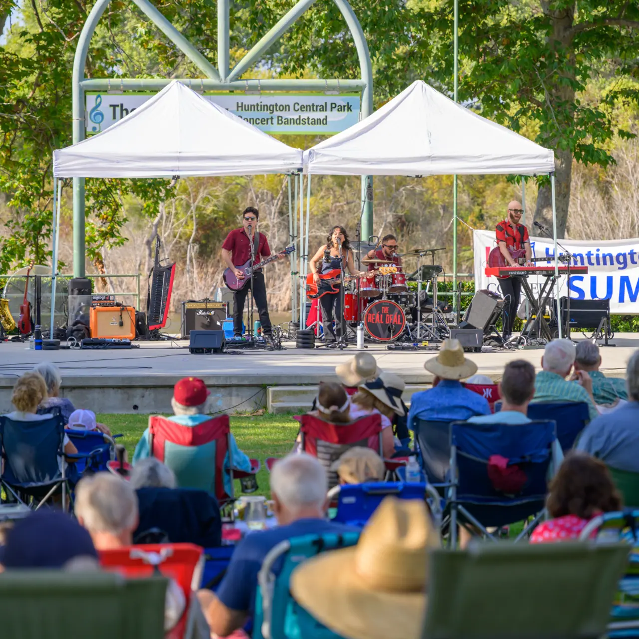 A live band performing on stage at Huntington Central Park during a summer event, with an audience seated in front.