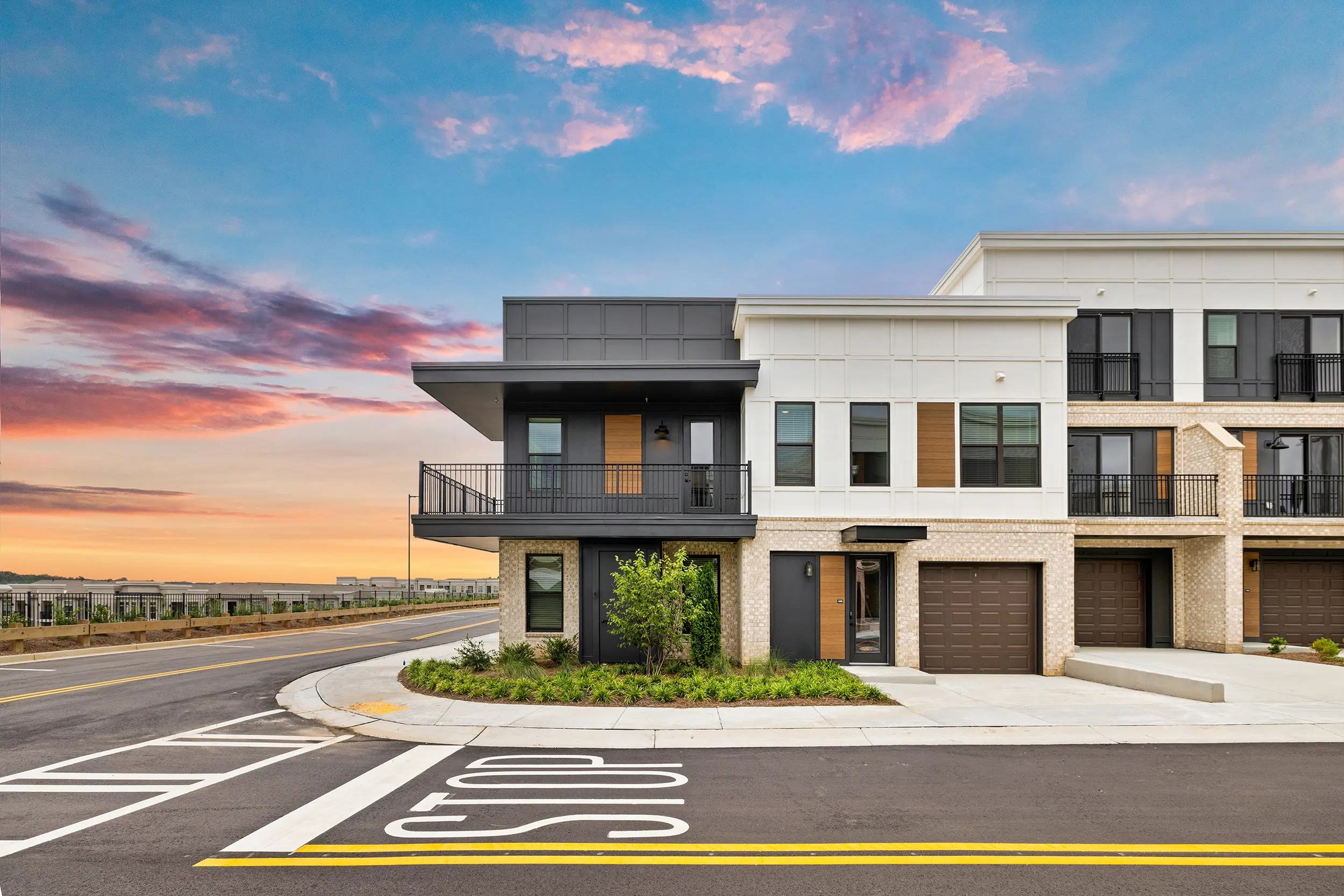 A modern building exterior with a two-toned facade—white and dark gray—featuring balconies and large windows, situated at a street corner with a dramatic sunset sky.