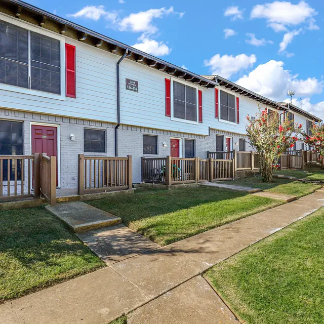 A view of a townhouse complex with two-story buildings. The buildings have red doors and windows, surrounded by grass and pathways. Trees and shrubbery are present in the foreground.