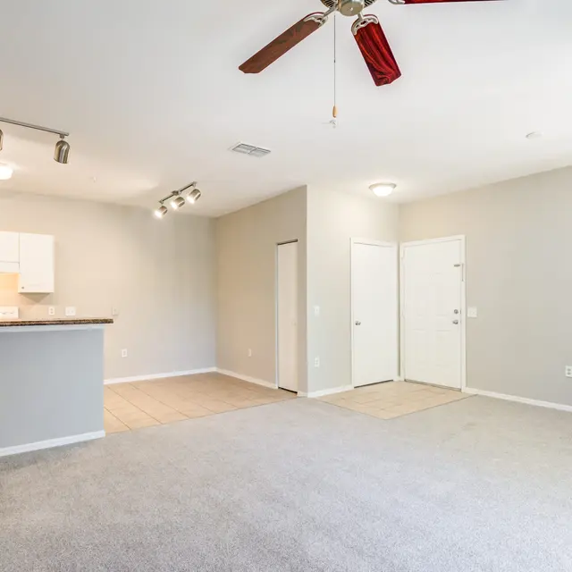 A spacious, empty living area with beige carpet and a ceiling fan. The kitchen area is in the background, featuring white cabinets and a tiled floor.