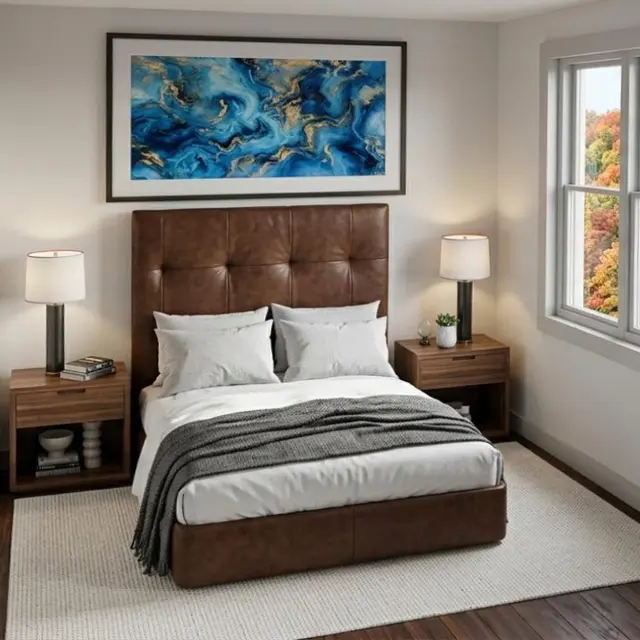 A modern bedroom featuring a brown upholstered bed with white linens, two bedside lamps, and decorative artwork above the bed. The room has windows with a view of colorful foliage outside.