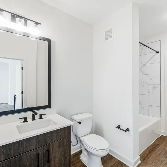 A modern bathroom featuring a dark wood vanity with a white countertop, a large mirror with a black frame, and a toilet. The shower area has a white tiled wall with a bathtub, and the floor is made of wood-look tile.