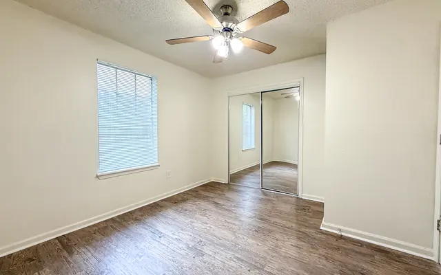 A spacious empty room with wooden flooring, a ceiling fan, and a large mirror closet. There is a window on the left allowing natural light to illuminate the space.