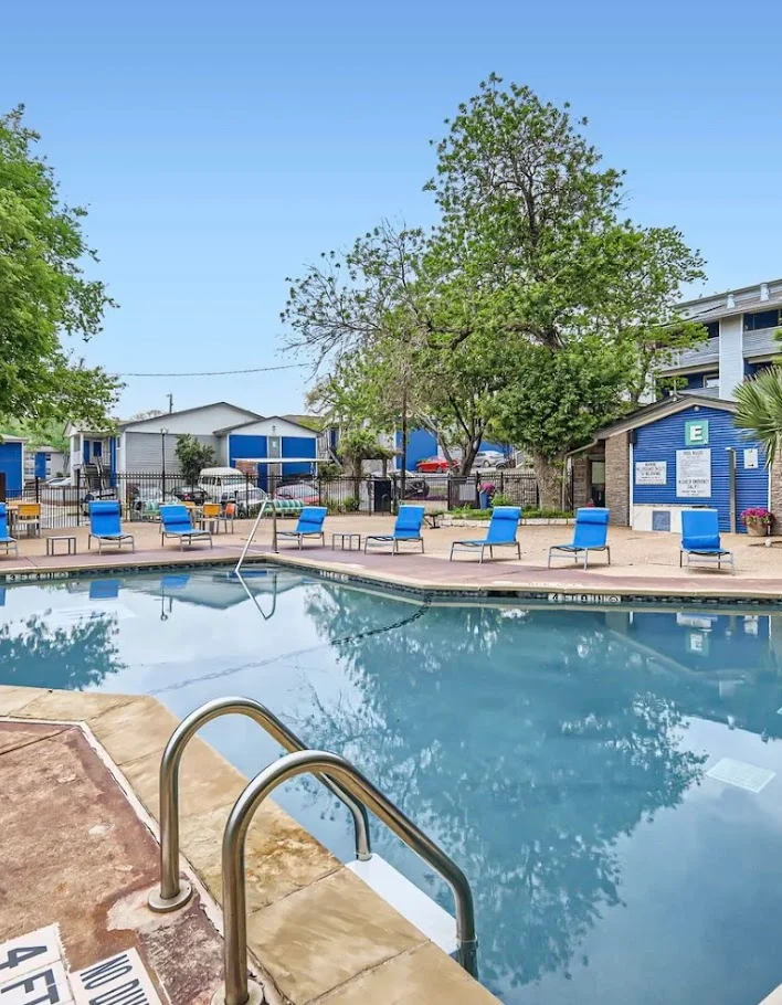 A swimming pool surrounded by lounge chairs and greenery, with blue buildings in the background.