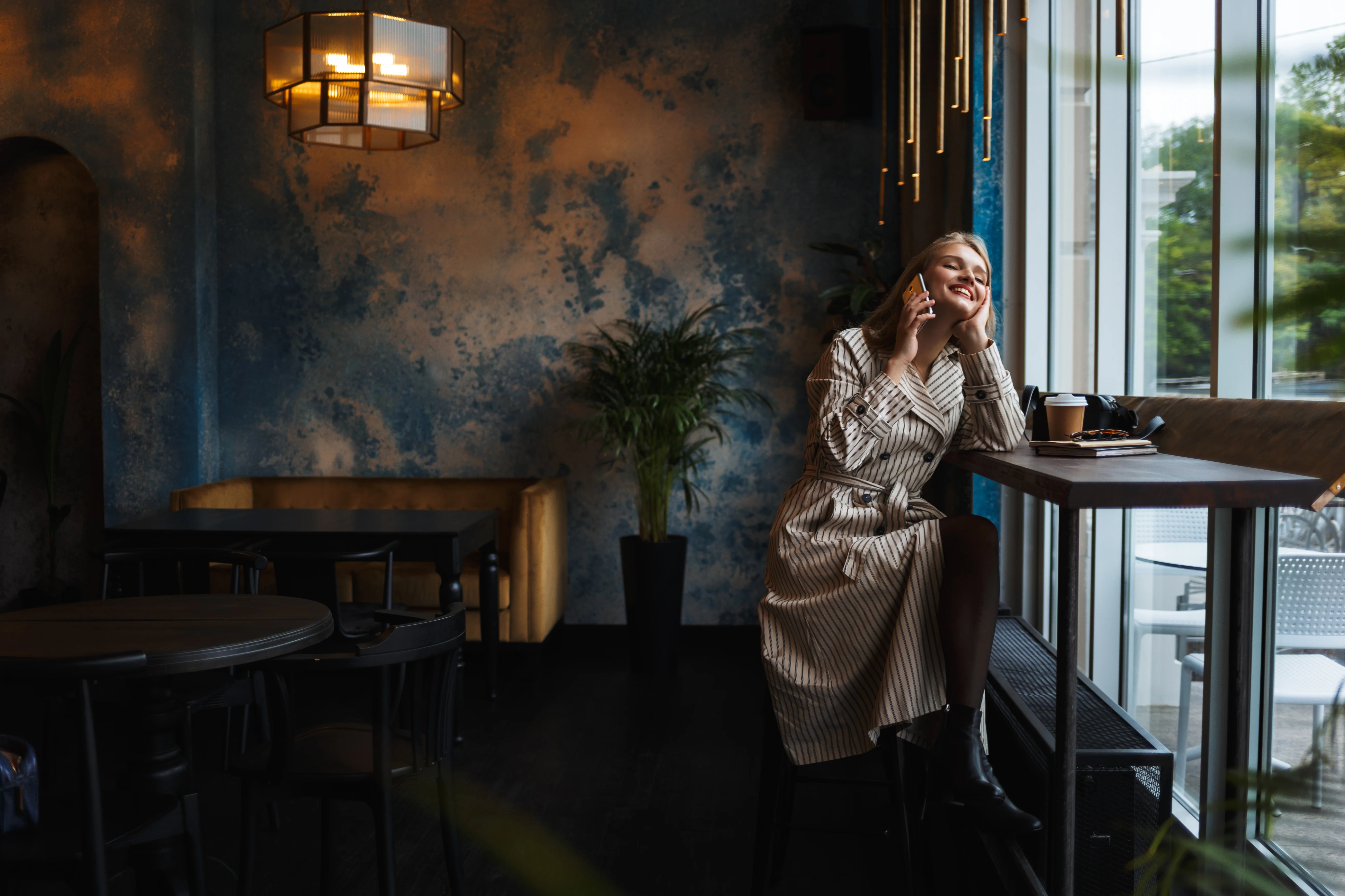Smiling Woman in a Cozy Cafe A woman in a striped dress smiling while sitting in a cafe, near a large window. The interior is stylish with a blue wall, plants, and modern lighting fixtures.