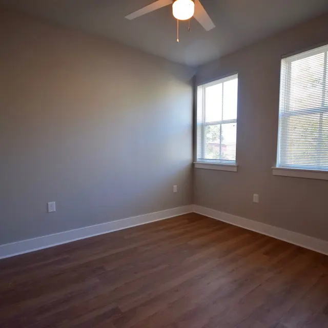 An empty room with light brown wooden flooring, two windows allowing natural light, and a ceiling fan.