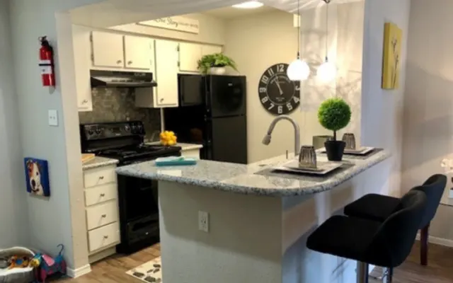 Modern Kitchen with Bar Seating A modern kitchen with a bar area featuring two black seats, a gray countertop, and a small green plant. The kitchen has white cabinets, a black refrigerator, and an oven, with decorative items on the counter and a large wall clock.