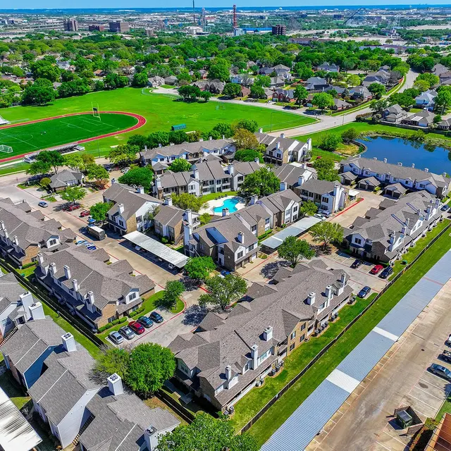 Aerial view of a residential apartment complex with a swimming pool, surrounded by green lawns, trees, and a large field in the background. A pond is visible in the right section of the image.