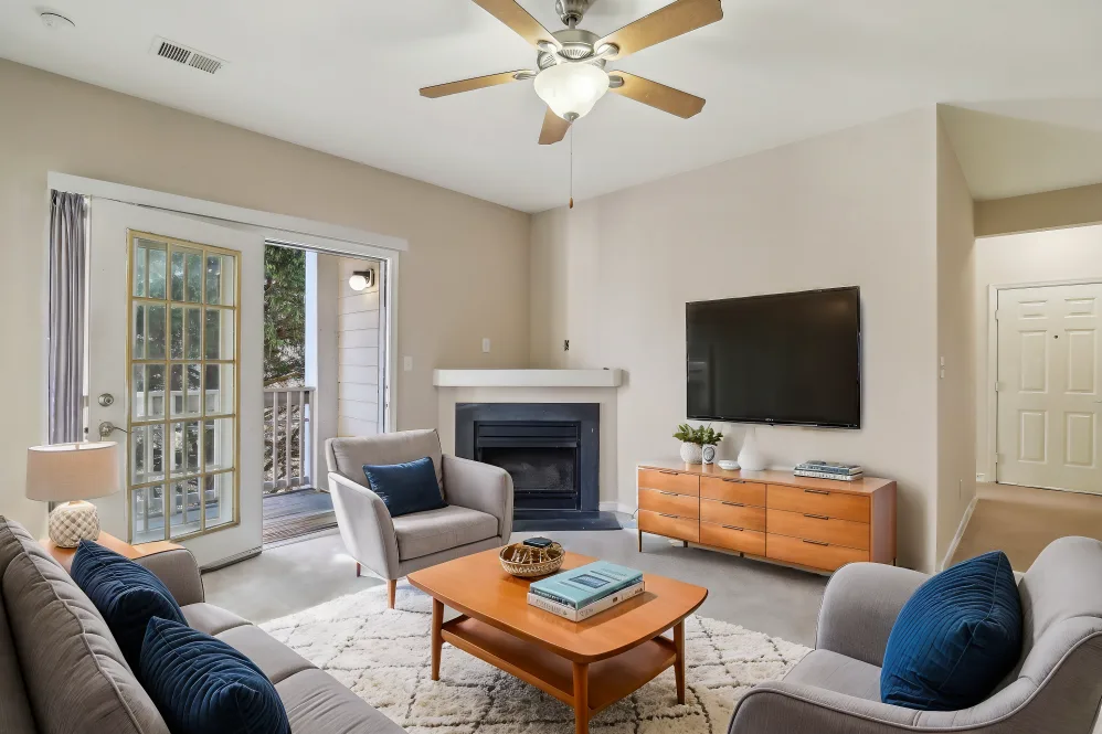 A modern living room featuring a cozy seating arrangement with two gray chairs and a sofa. A wooden coffee table sits in the center, adorned with books and decorative items. A flat-screen TV is mounted on the wall beside a fireplace. French doors lead to an outdoor space, while neutral-colored walls complement the overall decor.