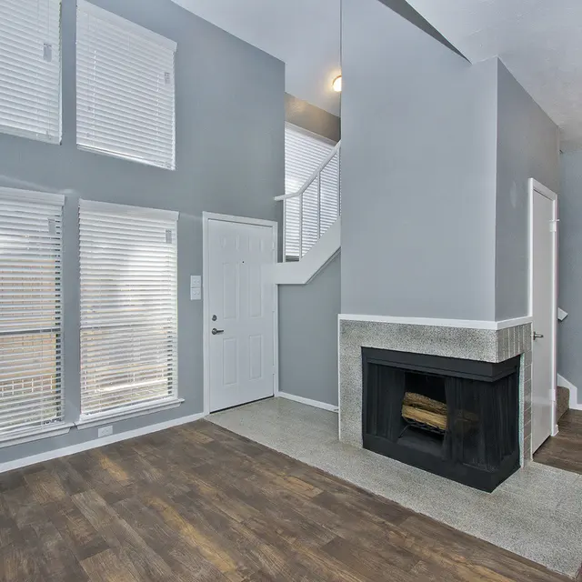 A modern living room featuring large windows with white blinds, a front door, and a gray wall with a small fireplace. The floor is wooden, and there is carpet near the fireplace.