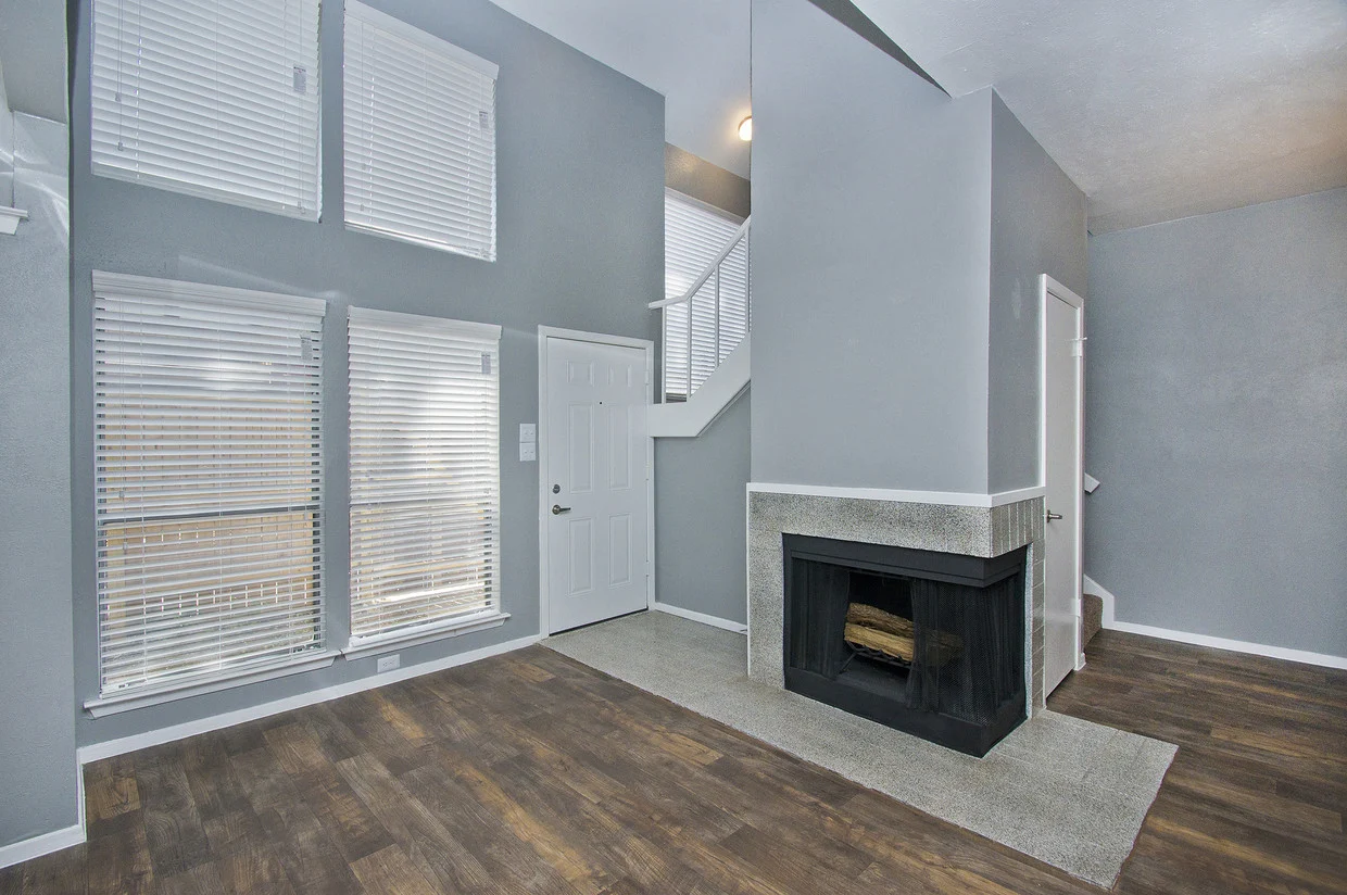 A modern living room featuring large windows with white blinds, a front door, and a gray wall with a small fireplace. The floor is wooden, and there is carpet near the fireplace.