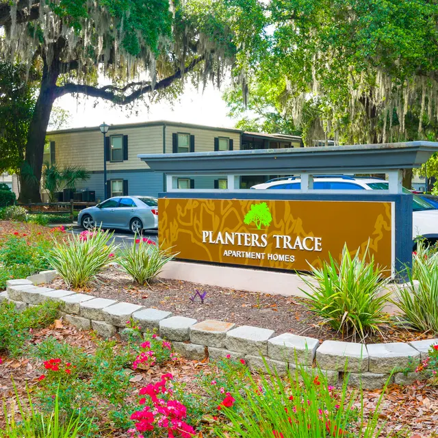 Sign for Planter's Trace Apartment Homes amidst landscaped greenery and flowering plants.