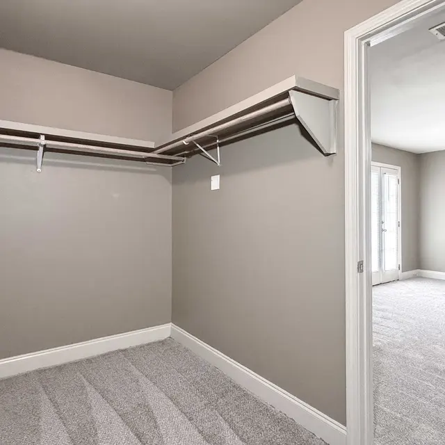 An empty closet with gray walls and carpeting, featuring two hanging rods. An open door leads to another room with carpet.