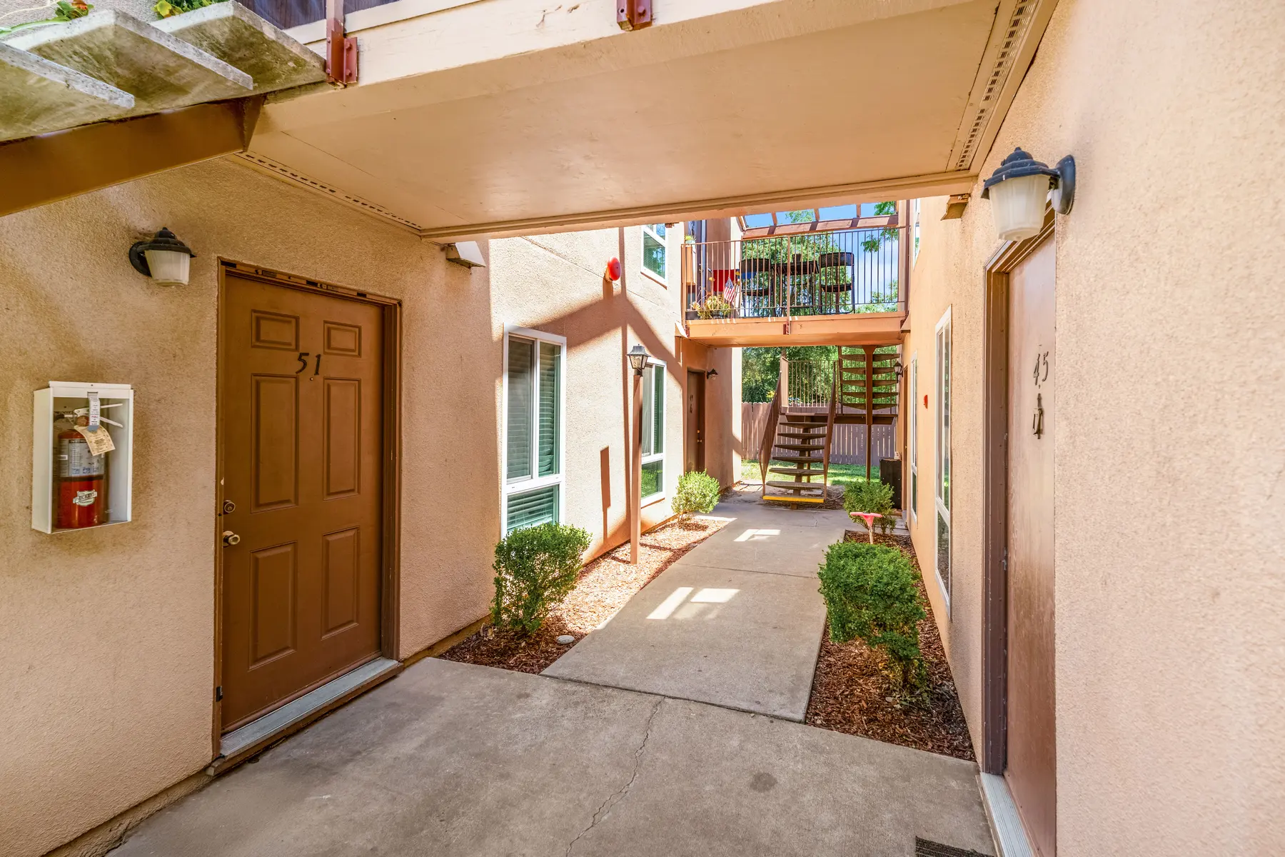 A view of an apartment courtyard showing a concrete pathway between two residential doors, with greenery and stairs leading to upper levels in the background.
