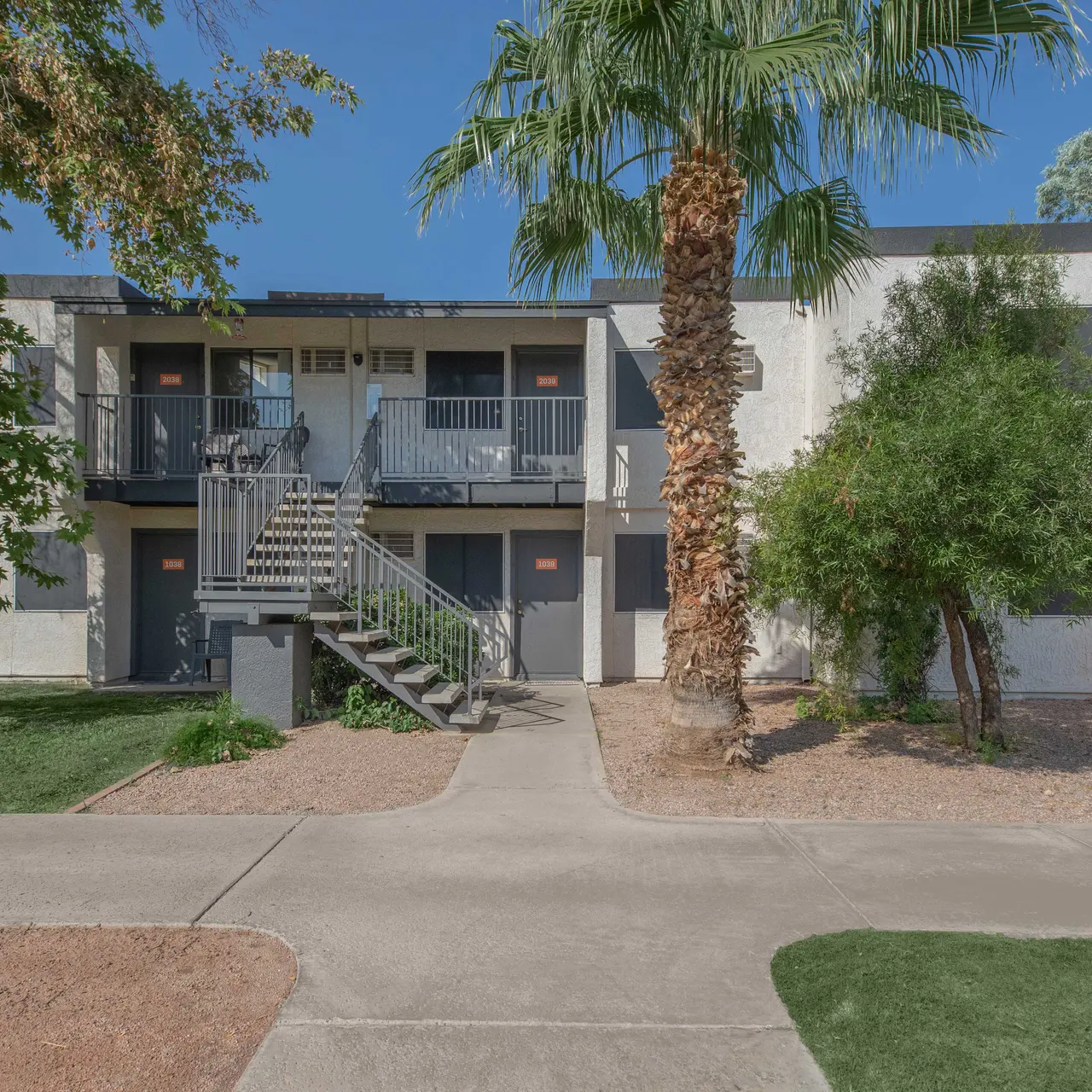 A view of a two-story apartment complex surrounded by palm trees and landscaping. The building features a staircase leading to the second floor and has balconies with railings.