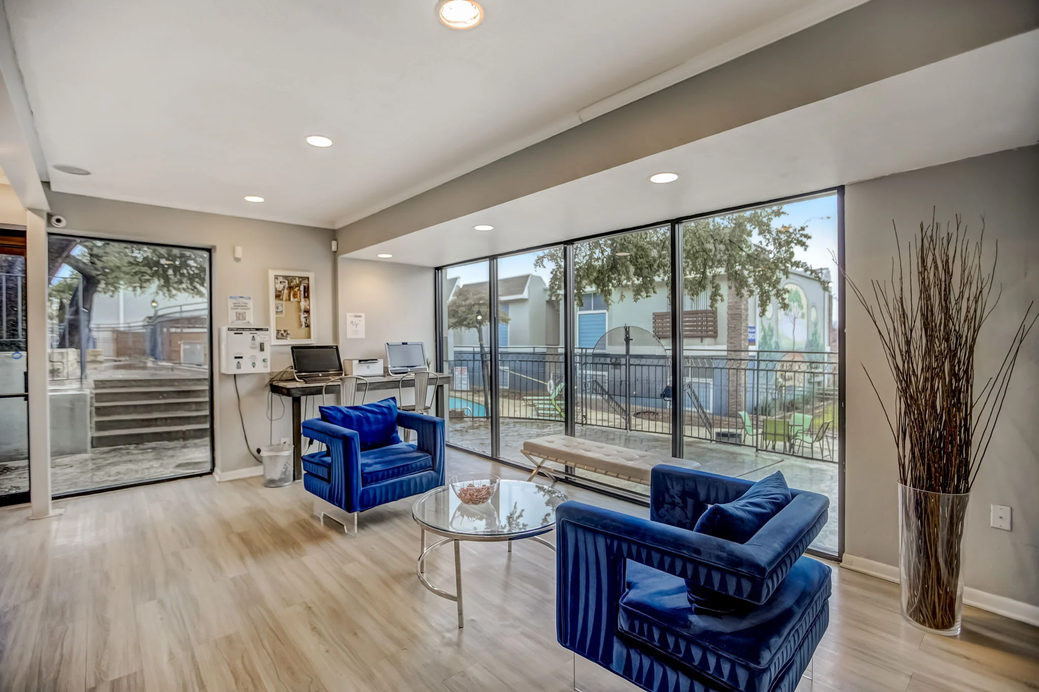 A modern lobby area with blue velvet chairs and a glass table, large windows offering a view of the outdoors, and light wooden flooring.