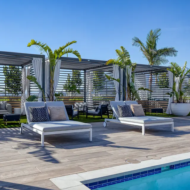 A luxurious rooftop lounge area with two white sun loungers beside a swimming pool, surrounded by potted palm trees and shaded seating arrangements under modern pergolas.