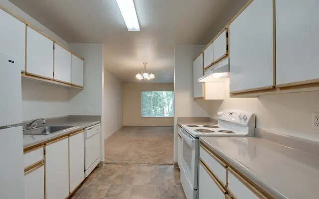 A clean kitchen with white cabinets and appliances, featuring a sink, oven, and a large window leading into a living area.