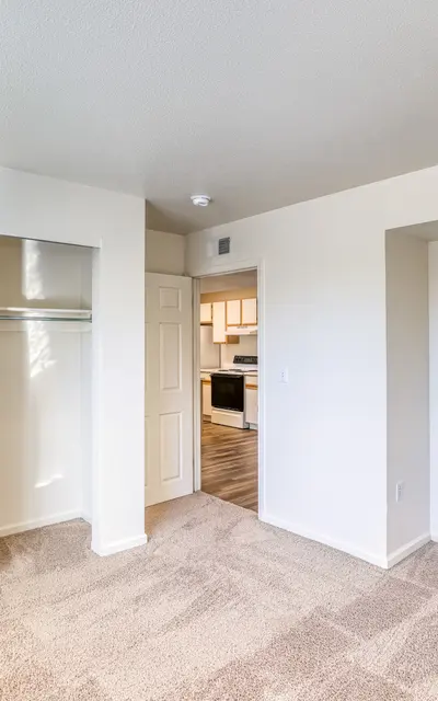 An empty room featuring beige carpet, white walls, and two doors—one leading to a closet and another leading to a kitchen. Natural light is coming in, illuminating the space.