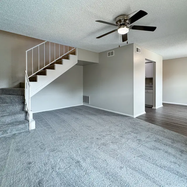 A spacious living room with carpeted flooring, featuring a set of stairs on the left leading to an upper level. The right side shows a kitchen area visible through an open doorway, and there is a ceiling fan overhead.