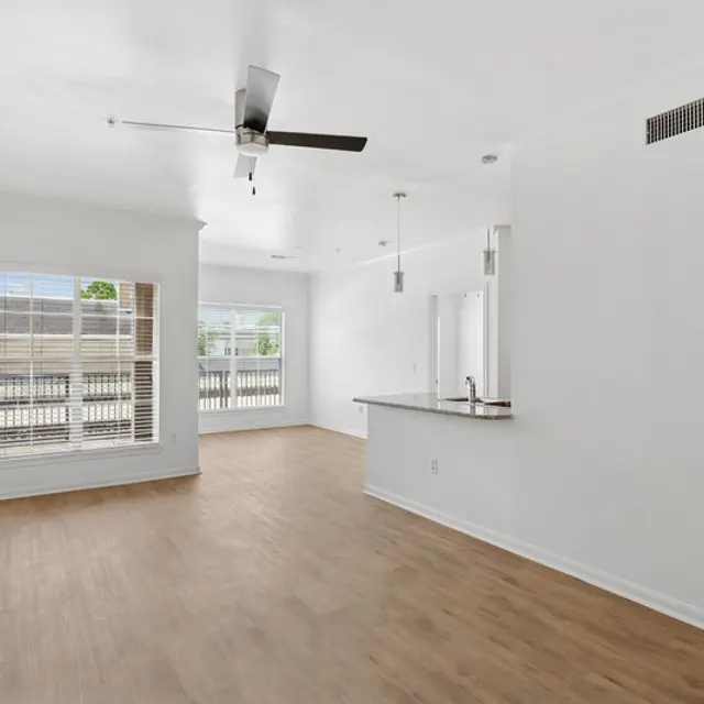 A spacious, modern apartment living room featuring light-colored walls, a ceiling fan, large windows with blinds, and a hardwood floor.