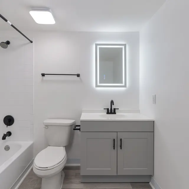 A modern bathroom featuring a bathtub with a black faucet, a white toilet, and a gray vanity with a black faucet. There is a round mirror with LED lighting above the vanity, and the walls are painted white with gray flooring.