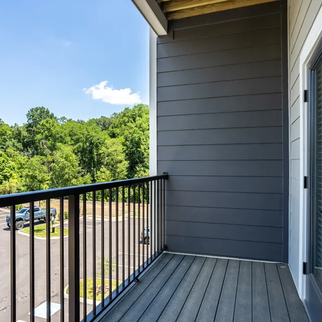 A balcony with a black railing overlooking a green landscape and a parking lot.