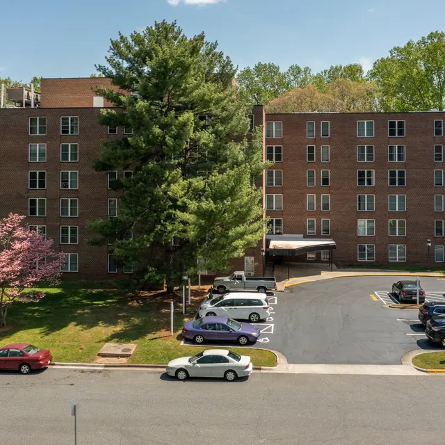 View of a multi-story brick apartment building surrounded by trees and a parking lot, featuring several parked cars, including a white car and a purple car. A pink flowering tree adds color to the scene.