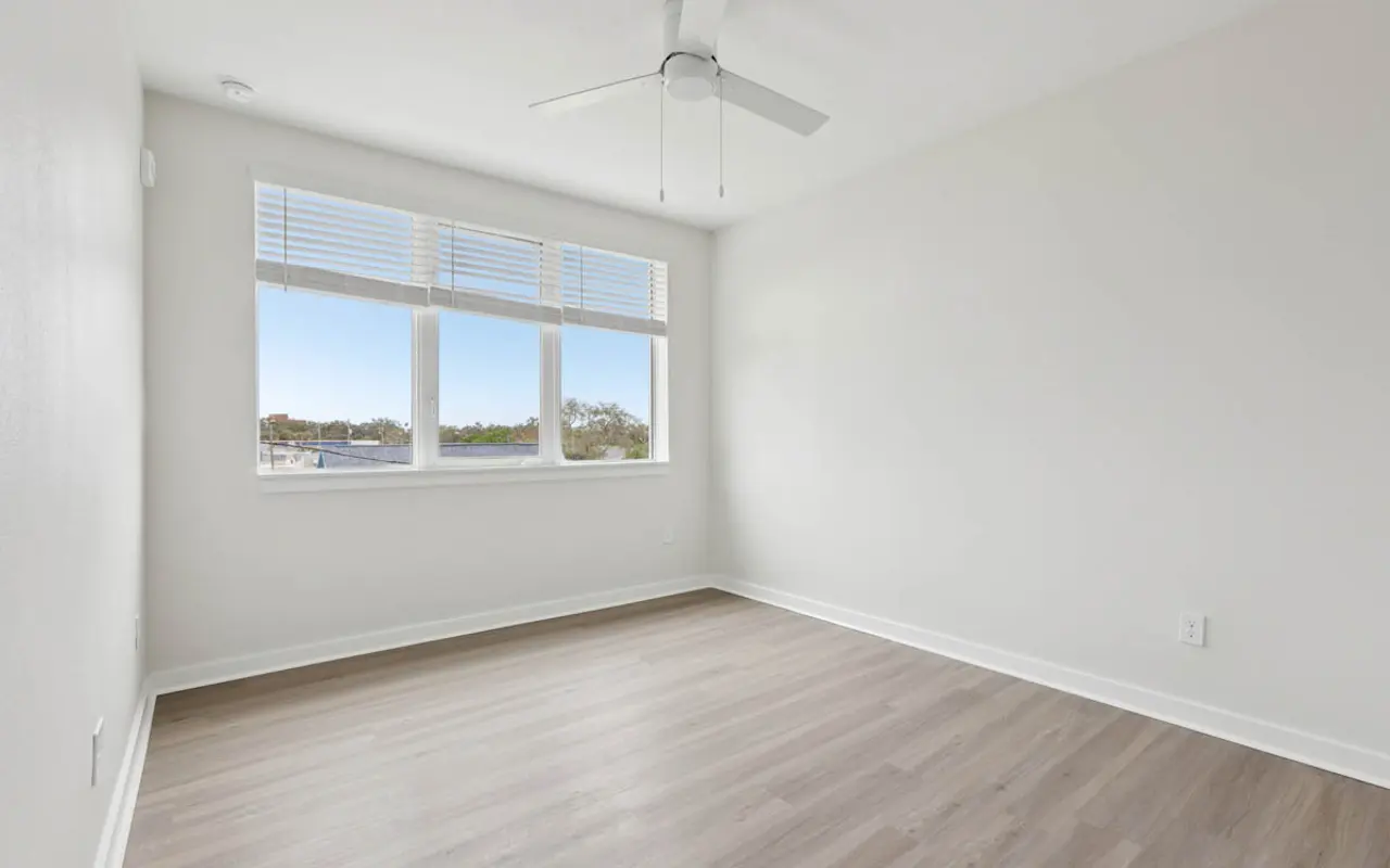 Bright and Spacious Empty Room A light and airy empty room with a ceiling fan and windows allowing natural light to enter. The room has light-colored walls and wooden flooring, creating a spacious feel.