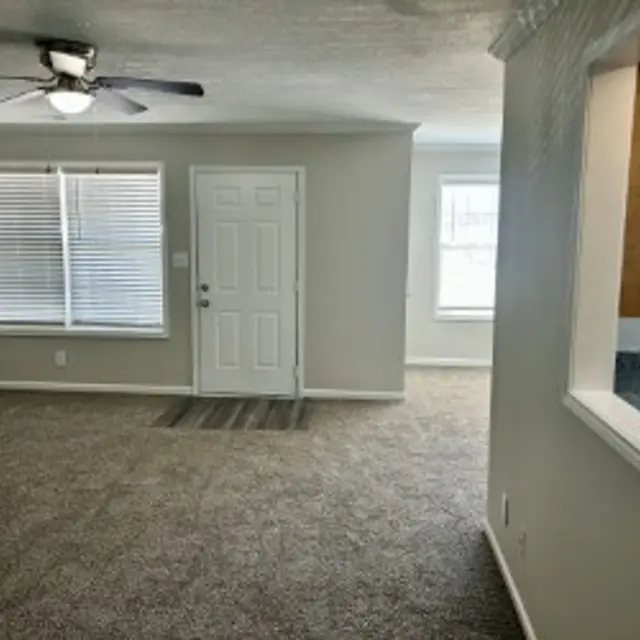 Spacious living room with beige walls and plush carpeting, featuring a ceiling fan and large windows for natural light.