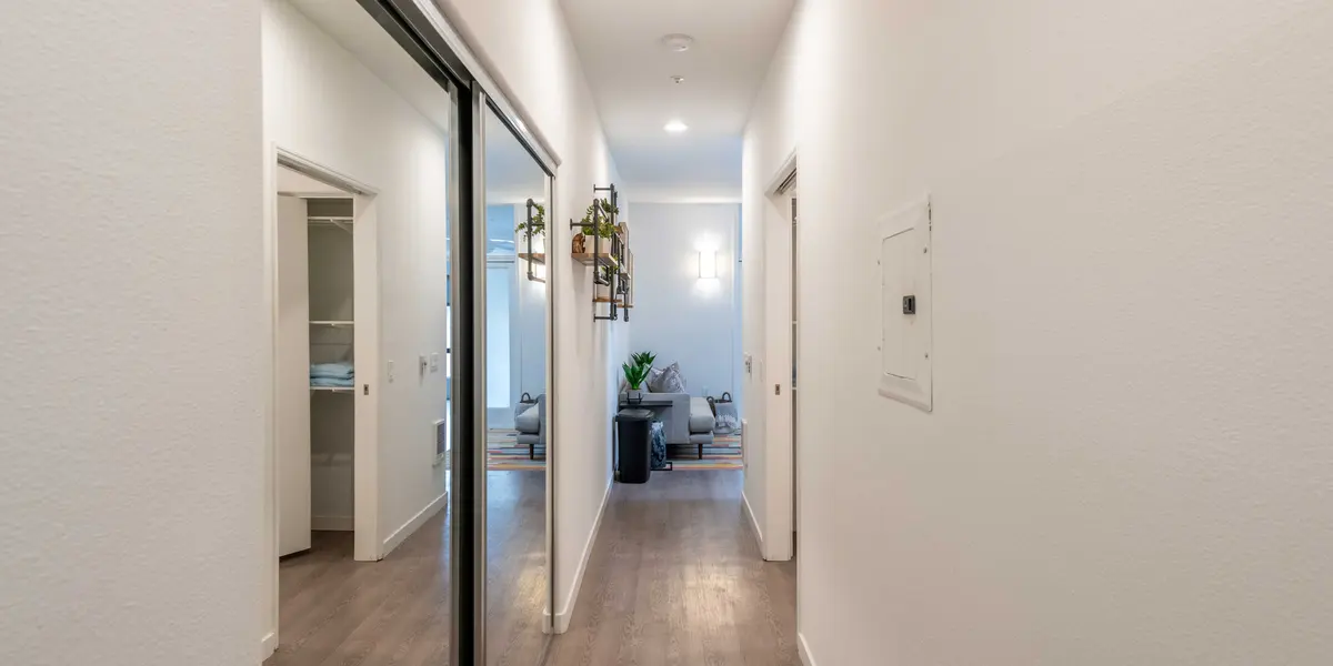A modern hallway with a sleek wooden floor and minimalistic decor. The walls are painted white, and there are doors on either side leading to other rooms. At the end of the hallway, a small seating area can be seen with a plant and stylish furniture.