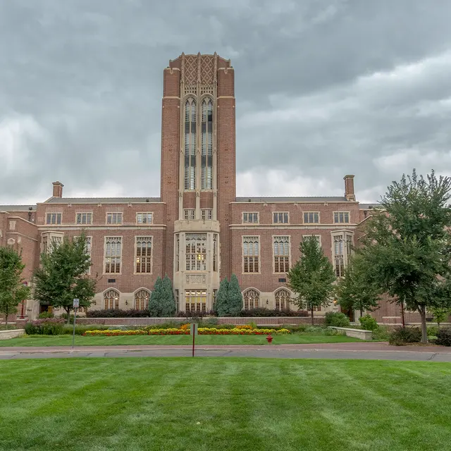 A large historic academic building with a tall tower, surrounded by greenery and a well-maintained lawn under a cloudy sky.