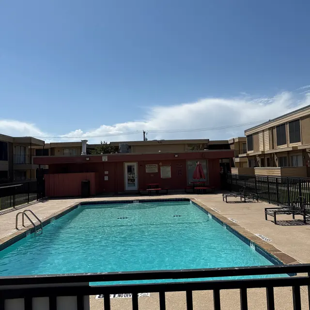 A swimming pool surrounded by a fence, with clear blue water under a sunny sky. Apartment buildings are visible in the background.