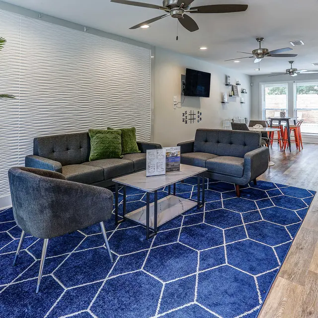 A modern living room featuring a grey couch and armchairs on a blue geometric rug. The space has a textured white wall, decorative plants, and a coffee table with brochures. There are two ceiling fans and large windows leading to an outdoor area.