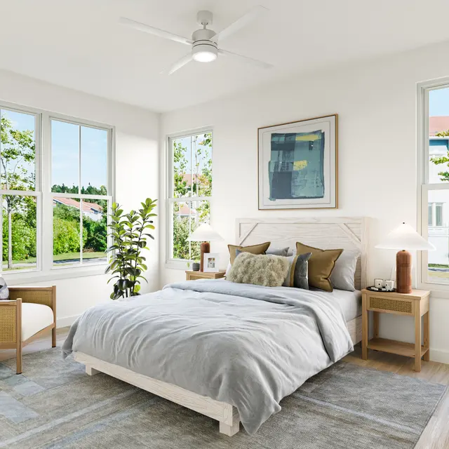 A modern bedroom featuring a comfortable bed with gray bedding, two bedside tables with lamps, a cozy armchair, potted plants, and multiple windows allowing natural light to fill the space.