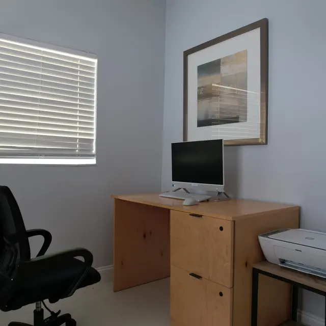 A minimalist home office space featuring a wooden desk with a computer, a black ergonomic chair, and a printer beside the desk. A window with blinds provides natural light, and a framed artwork hangs on the wall.