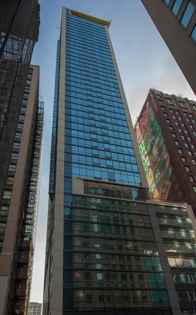 A tall modern skyscraper with glass windows, reflecting its surroundings, flanked by shorter buildings in an urban setting.