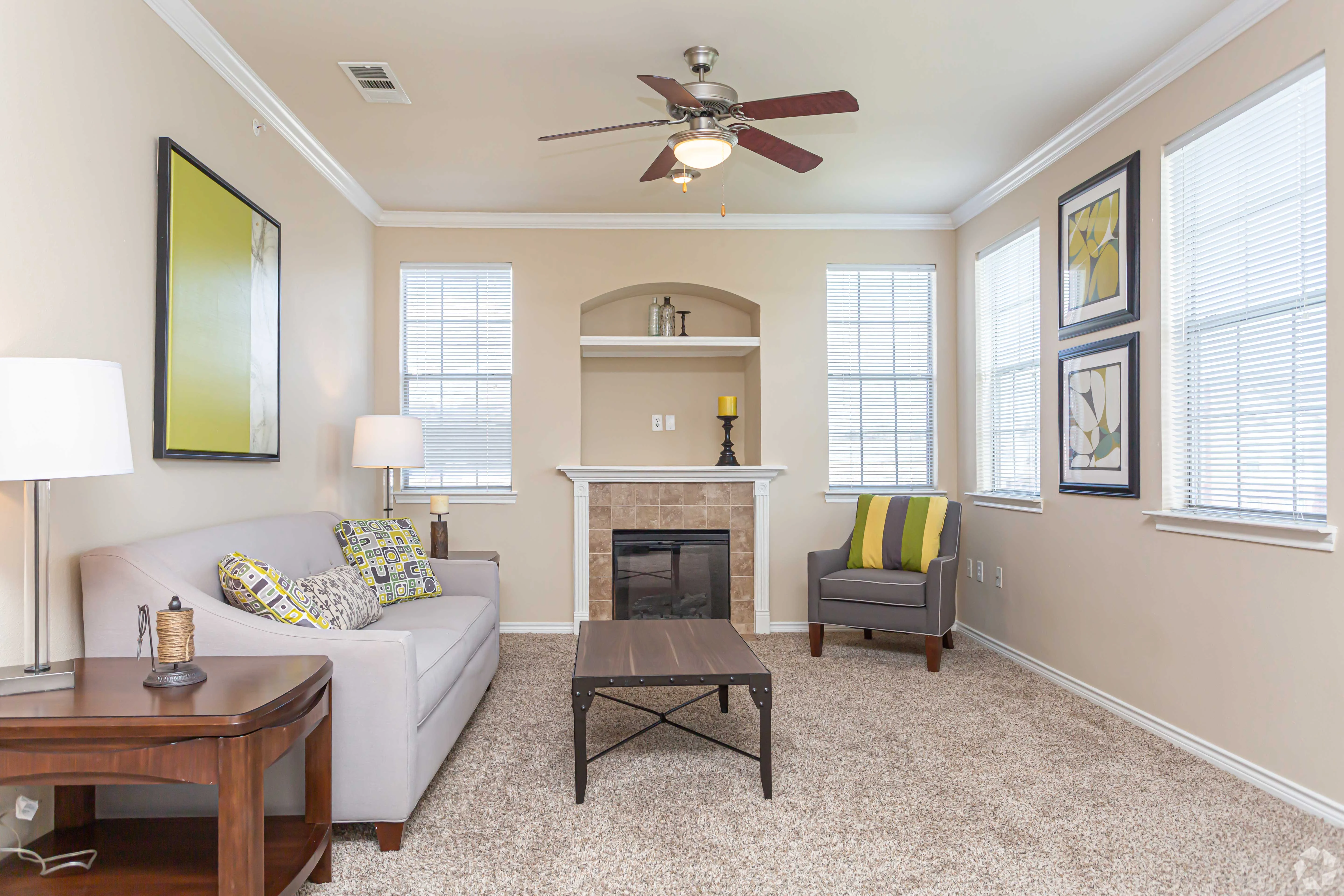 Cozy living room with a light gray sofa, decorative pillows, a wooden side table, and a fireplace. Large windows allow natural light, and artwork decorates the walls.