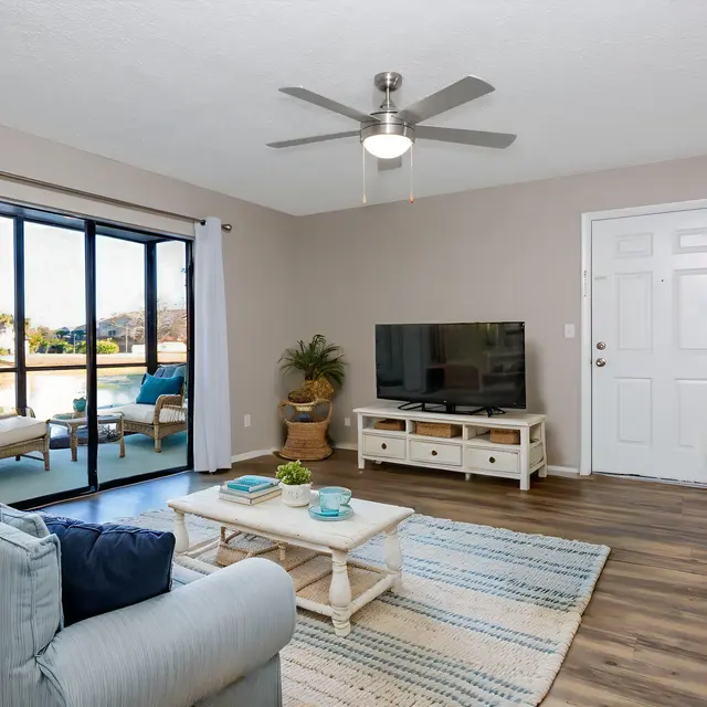 A cozy living room with a light blue couch, a coffee table, and a dining area visible in the foreground. A television is mounted on the wall facing the couch. In the background, there are large sliding glass doors that lead to an outdoor area with a view of trees and a landscape. The room has a ceiling fan and neutral-colored walls, creating a relaxed atmosphere.