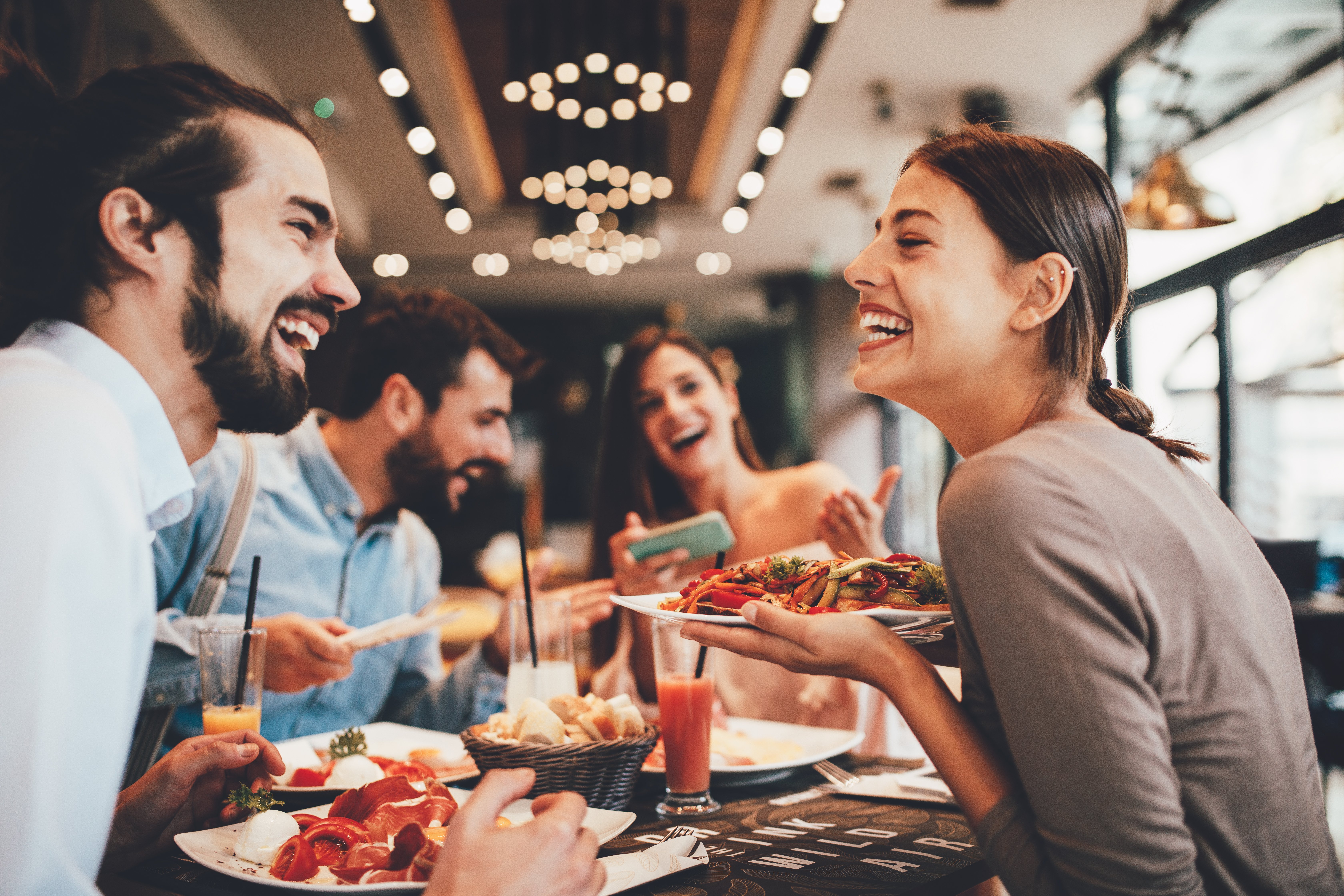 Friends Enjoying a Meal at a Restaurant A group of five friends enjoying a lively meal together at a restaurant. They are laughing and sharing food, with plates filled with colorful dishes in front of them. There are drinks on the table and a cheerful atmosphere.