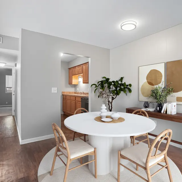 A modern dining area featuring a round table with a centerpiece and stylish chairs. The walls are painted in neutral tones, and natural light illuminates the space. A small plant adds a touch of greenery next to a decorative table with accessories. In the background, there is an open kitchen visible.