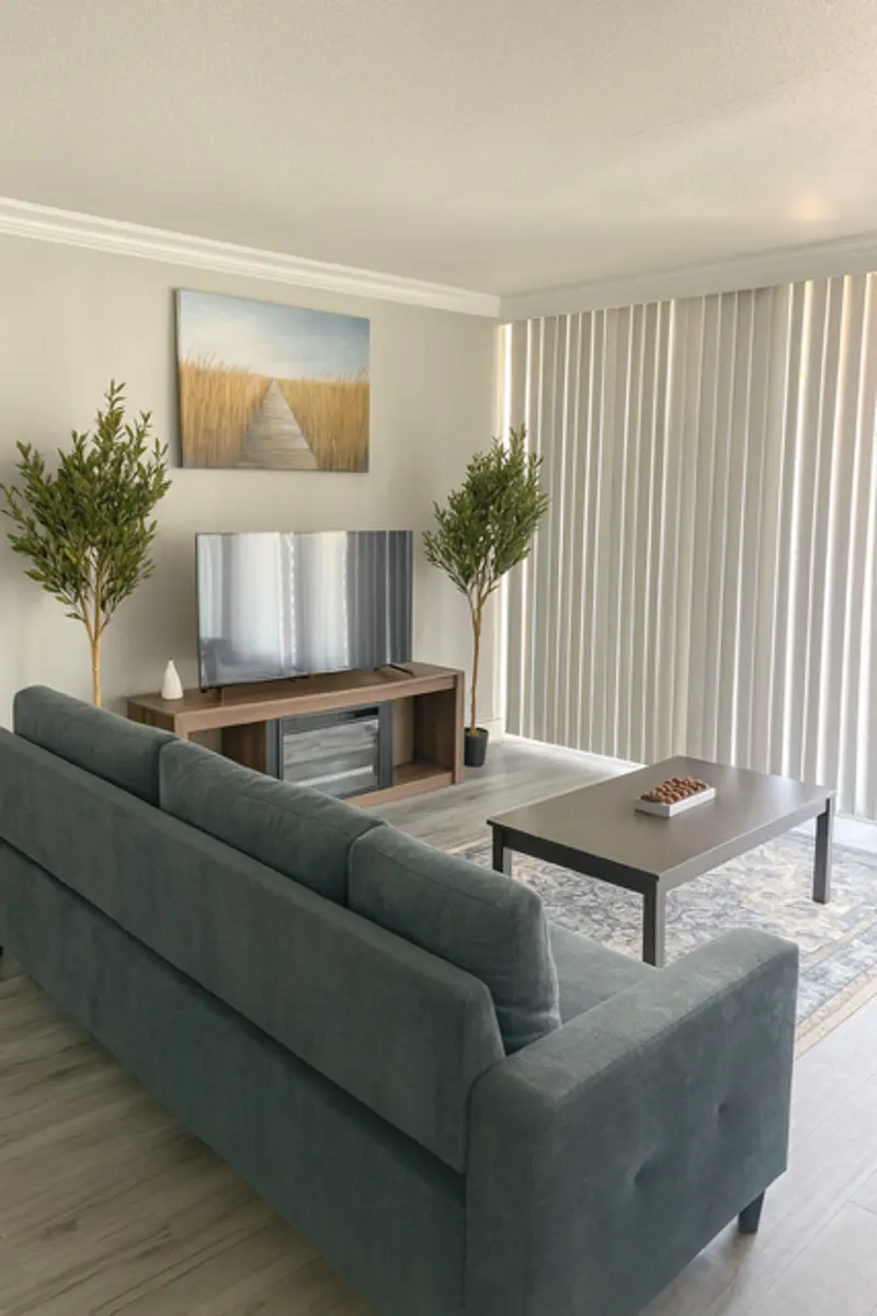 A modern living room setup featuring a teal couch, a wooden TV stand with a television, two indoor plants, a small coffee table with decorative items, and large windows with vertical blinds. A landscape painting is hanging on the wall above the TV.